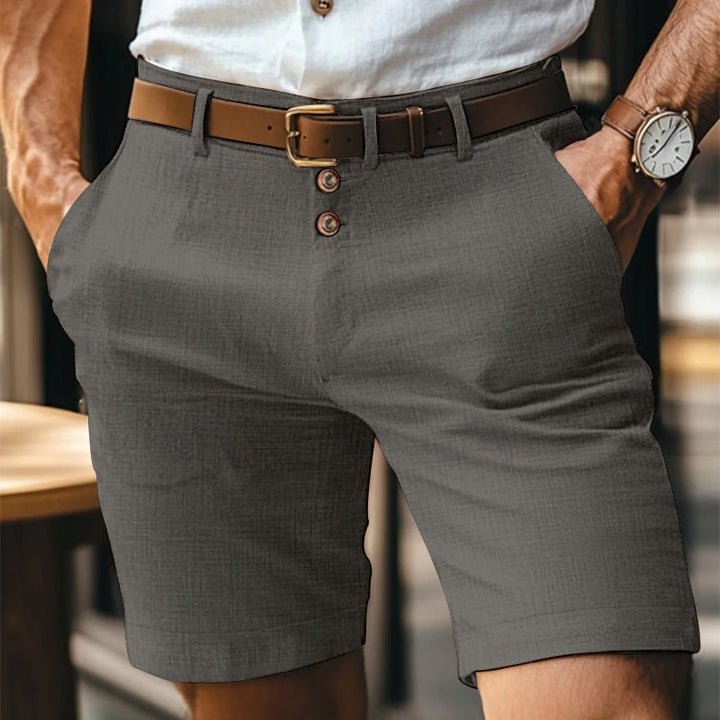 Uomo in pantaloncini grigi eleganti con cintura marrone e camicia bianca, orologio al polso. Moda estiva maschile, abbigliamento casual chic.