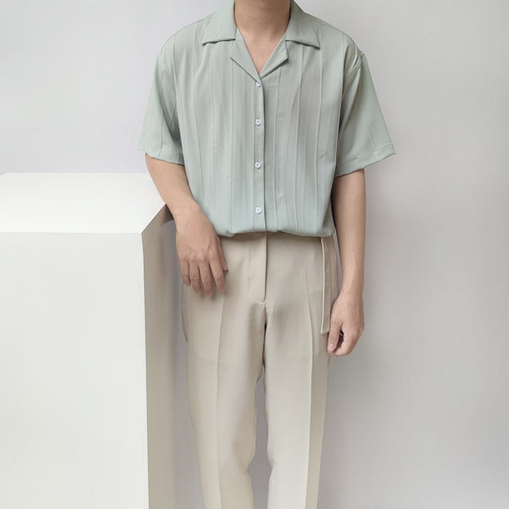 Uomo in camicia verde chiaro a righe verticali e pantaloni beige, posa accanto a un cubo bianco. Moda estiva, abbigliamento casual elegante.