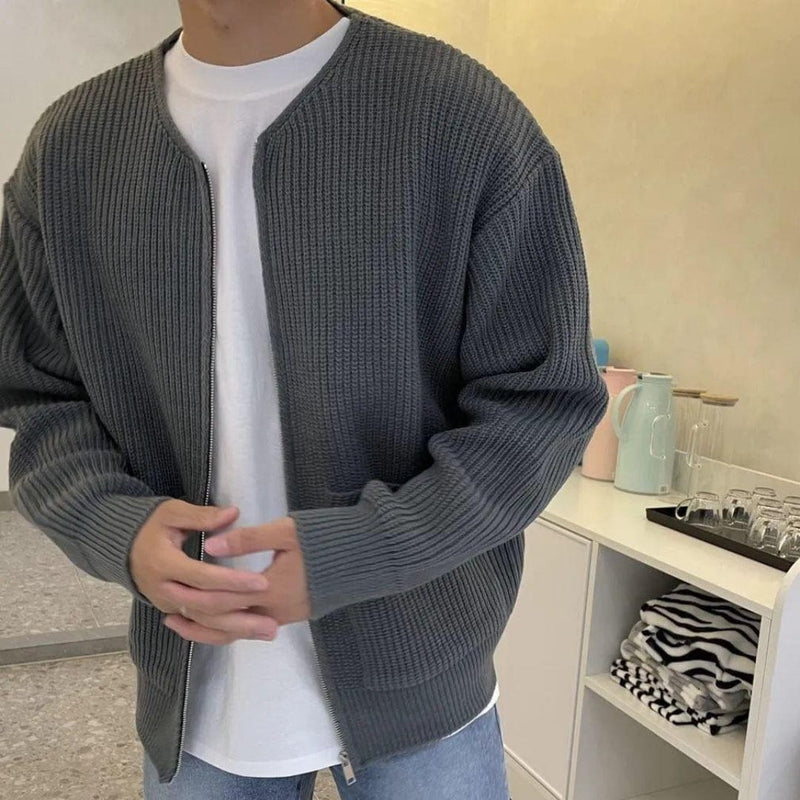 Uomo con cardigan grigio a maglia, t-shirt bianca e jeans. Sfondo con cucina moderna, caraffa azzurra e bicchieri. Moda casual autunnale.