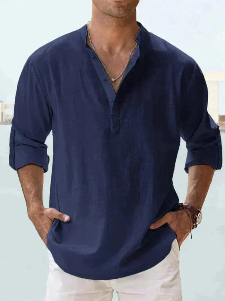 Camicia uomo blu scuro in lino, maniche lunghe arrotolate, collo alla coreana, abbinata a pantaloni bianchi. Stile casual elegante.