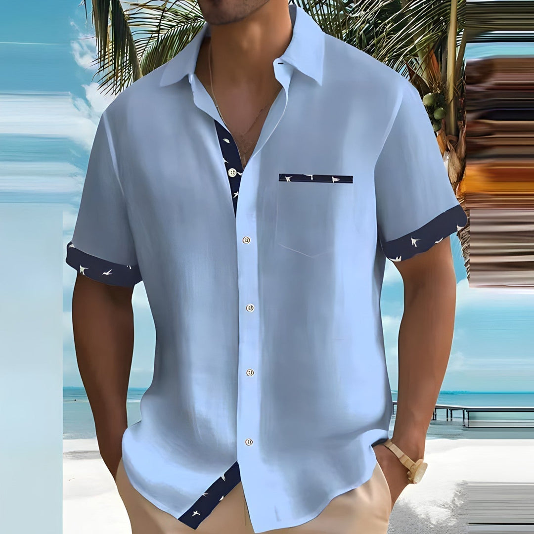Camicia estiva da uomo azzurra con dettagli a contrasto, indossata su spiaggia tropicale. Moda mare, abbigliamento casual, stile vacanza.