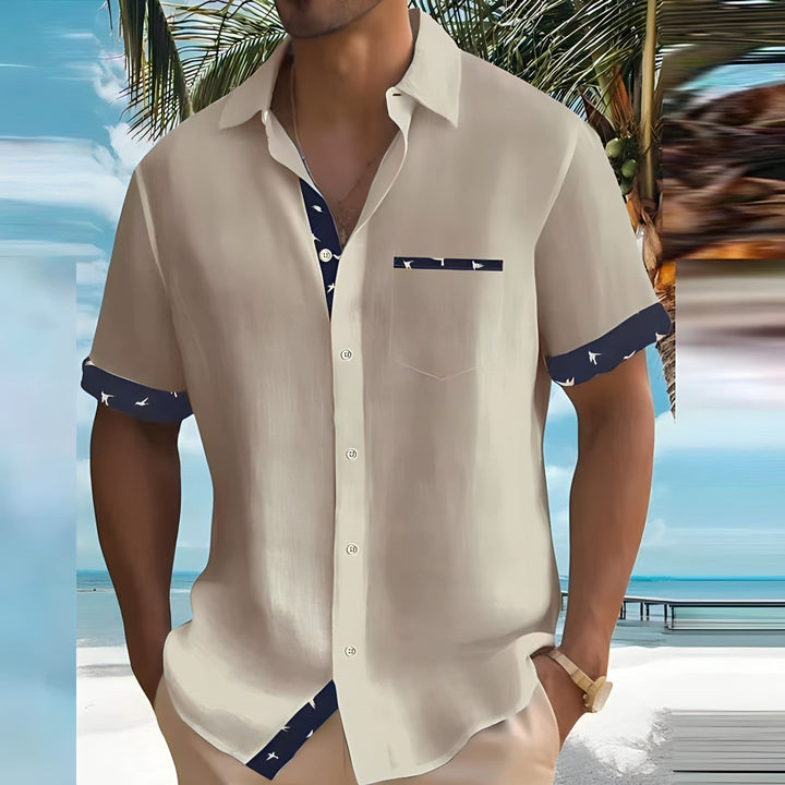 Camicia estiva da uomo beige con dettagli blu, indossata su una spiaggia tropicale. Moda casual, stile vacanza, palme sullo sfondo.