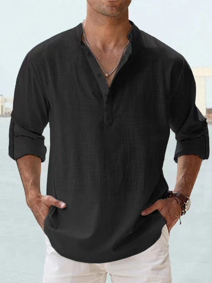 Camicia uomo nera in lino, maniche lunghe arrotolate, collo alla coreana, abbinata a pantaloni bianchi. Stile casual elegante.