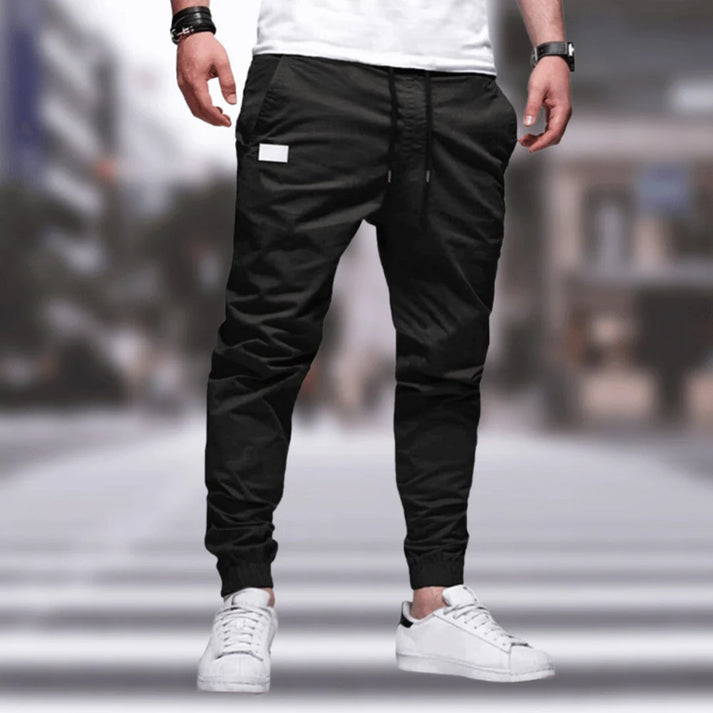 Uomo in pantaloni jogger neri e scarpe da ginnastica bianche su sfondo urbano sfocato. Moda casual maschile, streetwear moderno.