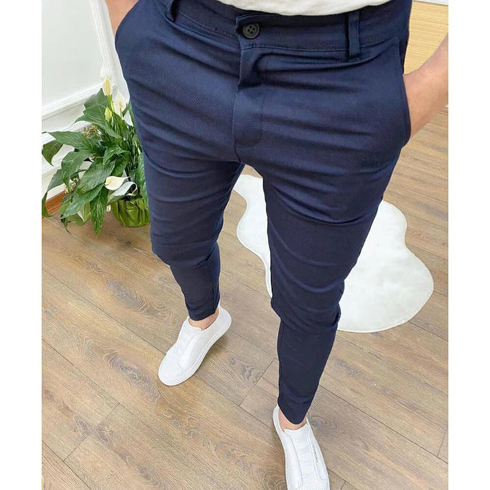 Pantaloni eleganti blu scuro da uomo, abbinati a scarpe bianche casual, su pavimento in legno con pianta verde sullo sfondo.