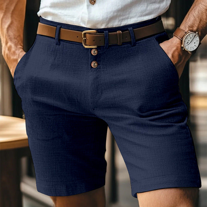 Uomo in camicia bianca e pantaloncini blu eleganti, con cintura marrone e orologio da polso, in un ambiente urbano. Moda estiva maschile.