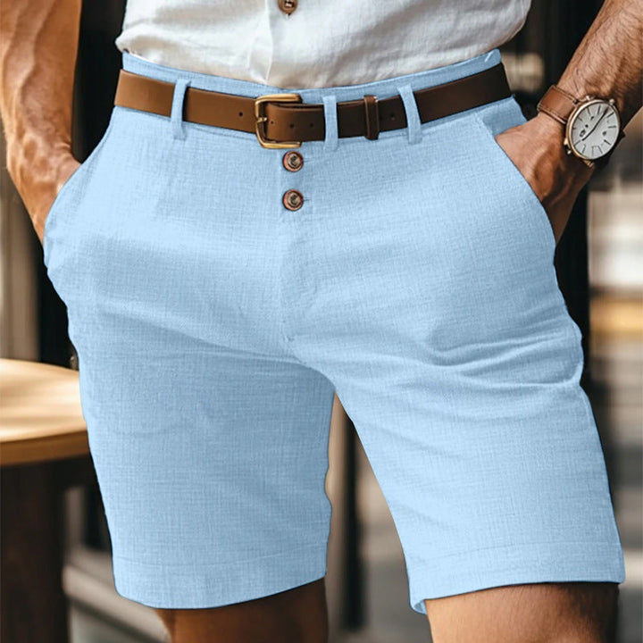 Uomo in pantaloncini azzurri eleganti, camicia bianca, cintura marrone, orologio al polso. Moda estiva maschile, abbigliamento casual chic.