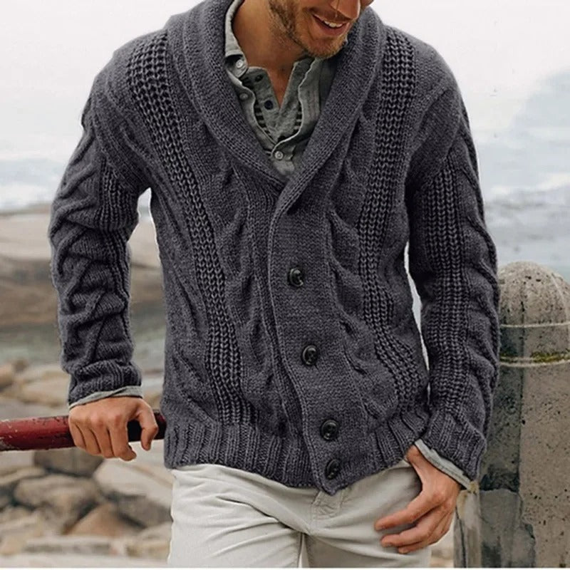Uomo con cardigan grigio a maglia grossa, bottoni grandi, su sfondo marino. Moda invernale, abbigliamento caldo, stile casual elegante.