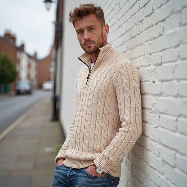 Uomo con maglione a trecce beige, appoggiato a un muro di mattoni bianchi, strada urbana sullo sfondo. Moda autunnale, stile casual elegante.